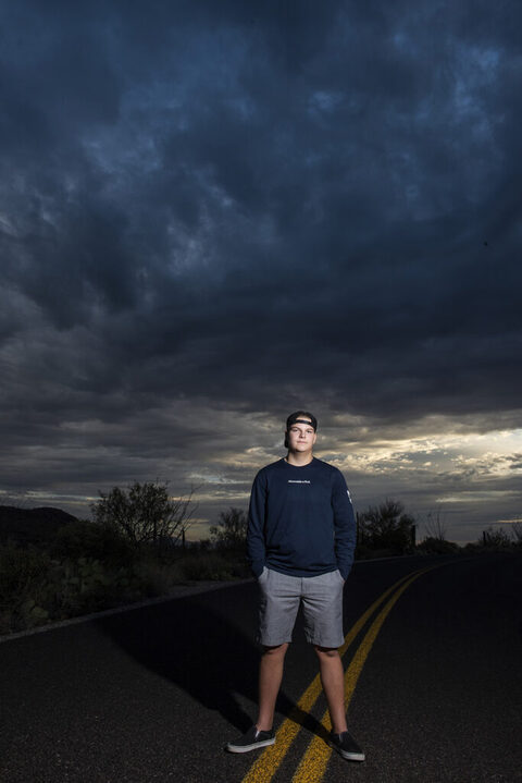 bot standing in the middle of a road on a yellow line wearing a hat backwards with dramatic skies for his tucson senior pictures for guys